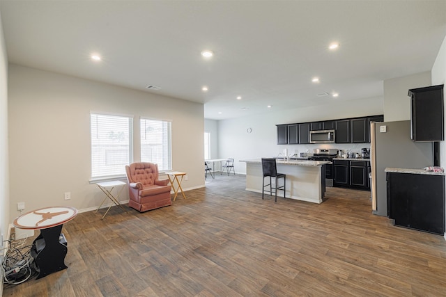 kitchen with a center island with sink, wood finished floors, recessed lighting, appliances with stainless steel finishes, and dark cabinets