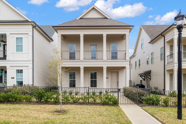 neoclassical / greek revival house featuring a front yard, a balcony, fence, roof with shingles, and stucco siding