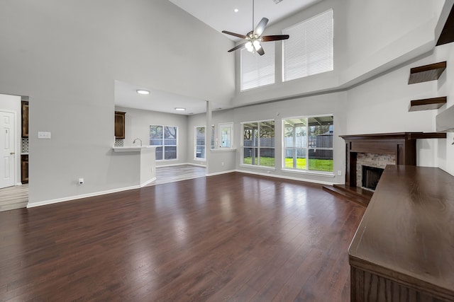 unfurnished living room with a ceiling fan, a high ceiling, a stone fireplace, baseboards, and dark wood-style flooring