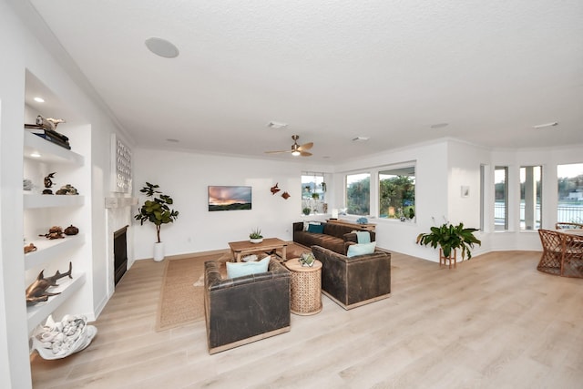 living room with light wood finished floors, built in features, ornamental molding, a fireplace, and a ceiling fan