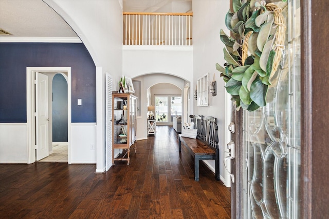 foyer entrance with wood finished floors, arched walkways, ornamental molding, a towering ceiling, and wainscoting