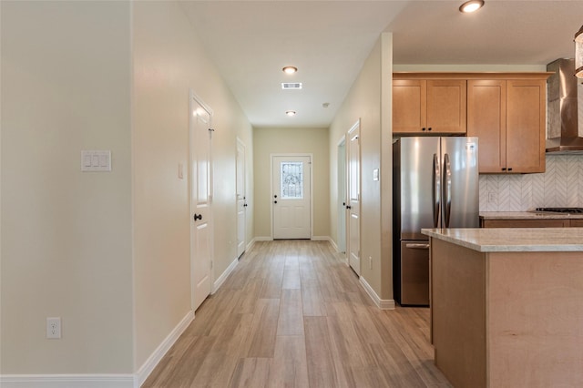 kitchen featuring wall chimney range hood, light countertops, decorative backsplash, freestanding refrigerator, and light wood-style floors