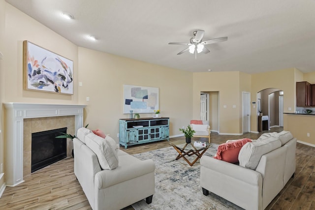 living area featuring wood finished floors, baseboards, arched walkways, a tile fireplace, and ceiling fan