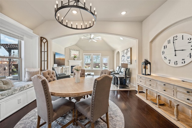 dining room with ceiling fan with notable chandelier, arched walkways, a fireplace, lofted ceiling, and dark wood-style flooring