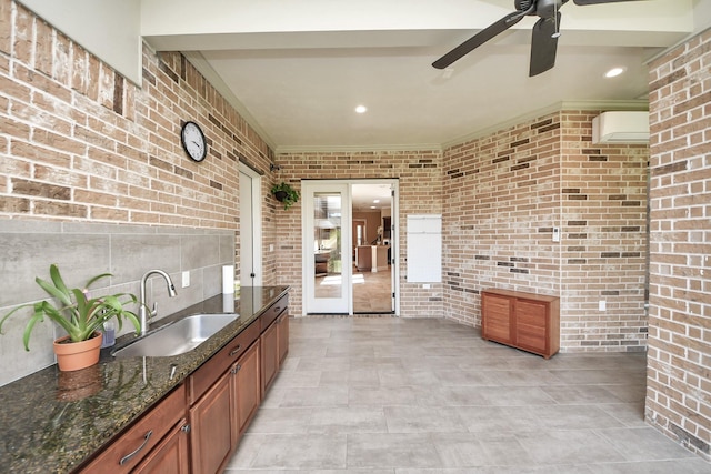 kitchen featuring a ceiling fan, a sink, dark stone counters, brick wall, and brown cabinetry