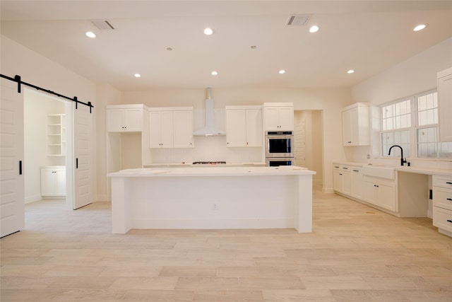 kitchen with a barn door, wall chimney exhaust hood, light wood-type flooring, and stainless steel double oven