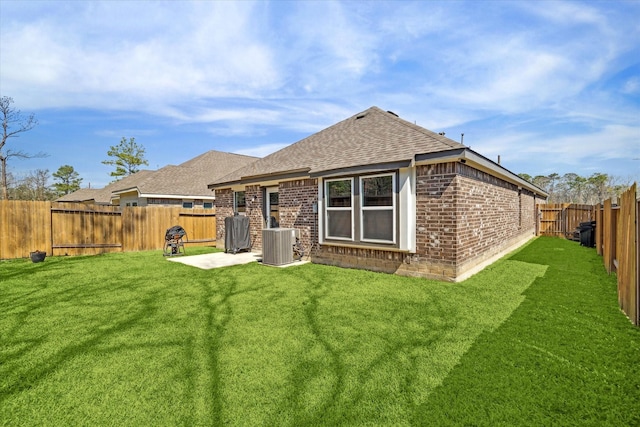 rear view of property with a fenced backyard, a yard, roof with shingles, brick siding, and central AC unit