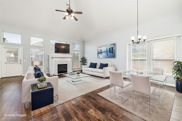 living area featuring wood finished floors, baseboards, high vaulted ceiling, a tiled fireplace, and ceiling fan with notable chandelier