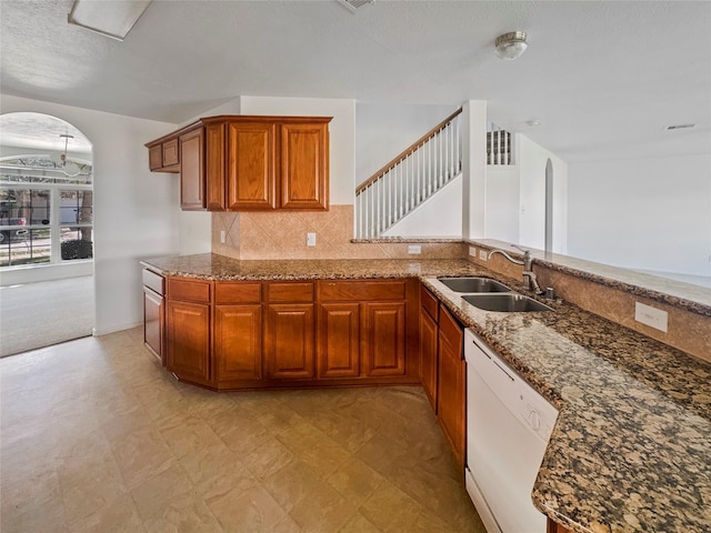 kitchen featuring dark stone countertops, brown cabinetry, arched walkways, a sink, and dishwasher