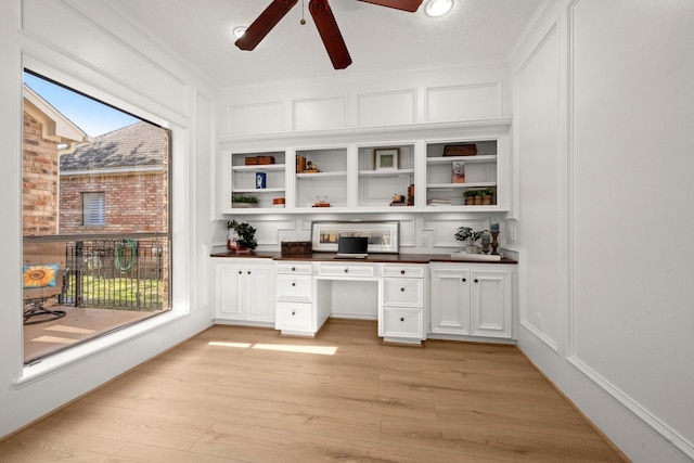 unfurnished office with light wood-style floors, a ceiling fan, built in desk, and a decorative wall