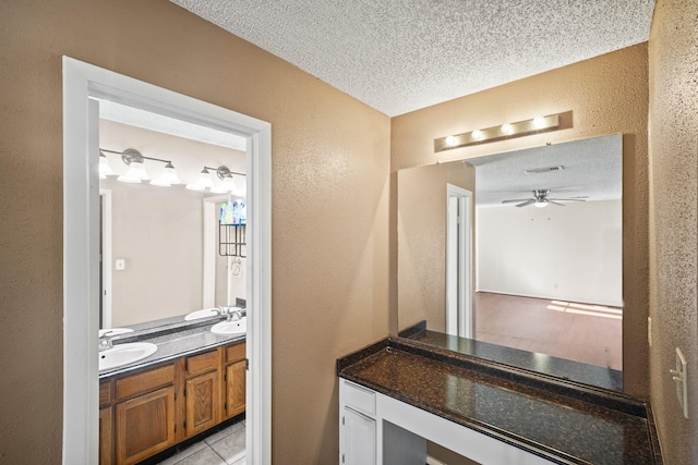 bathroom featuring ceiling fan, double vanity, a textured wall, a textured ceiling, and a sink