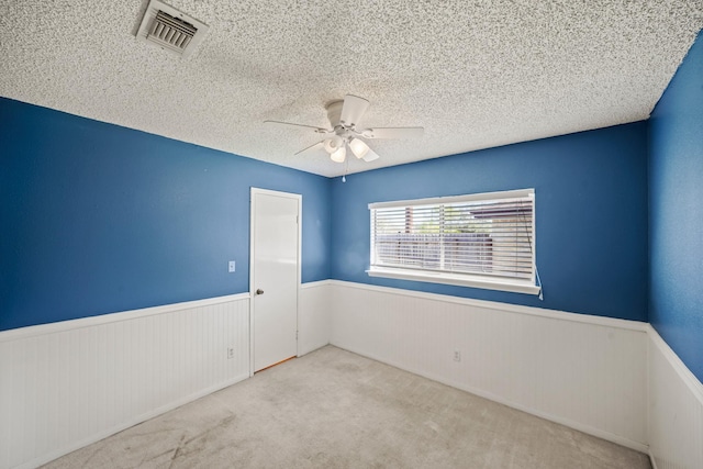 empty room featuring visible vents, a textured ceiling, carpet flooring, wainscoting, and ceiling fan