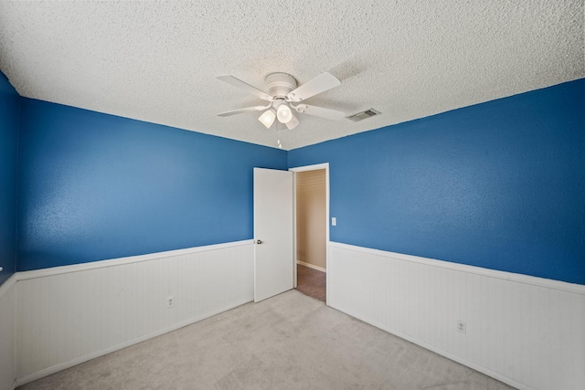 carpeted spare room with ceiling fan, visible vents, a textured ceiling, and a wainscoted wall