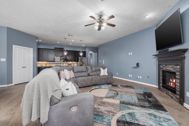 living area with baseboards, ceiling fan, a stone fireplace, light wood-style floors, and a textured ceiling