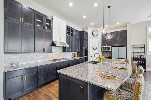 kitchen featuring under cabinet range hood, a sink, backsplash, dark wood-style floors, and built in appliances