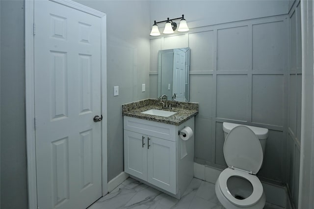 half bath featuring a decorative wall, marble finish floor, toilet, and vanity