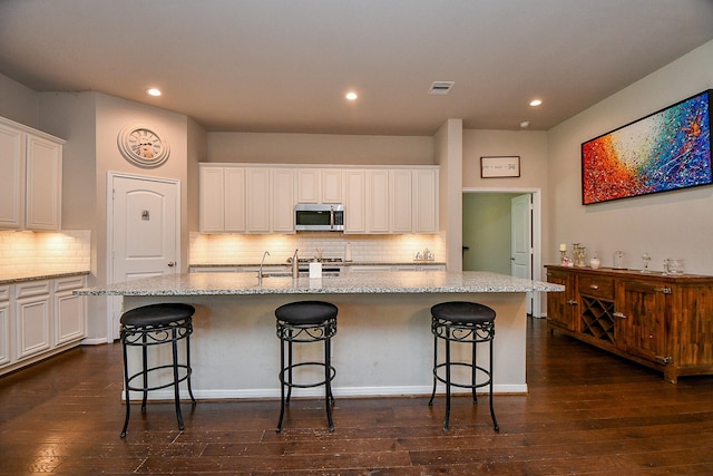 kitchen featuring stainless steel microwave, white cabinets, an island with sink, and dark wood-style flooring