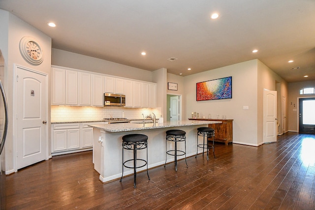 kitchen featuring stainless steel microwave, tasteful backsplash, an island with sink, and dark wood-style floors