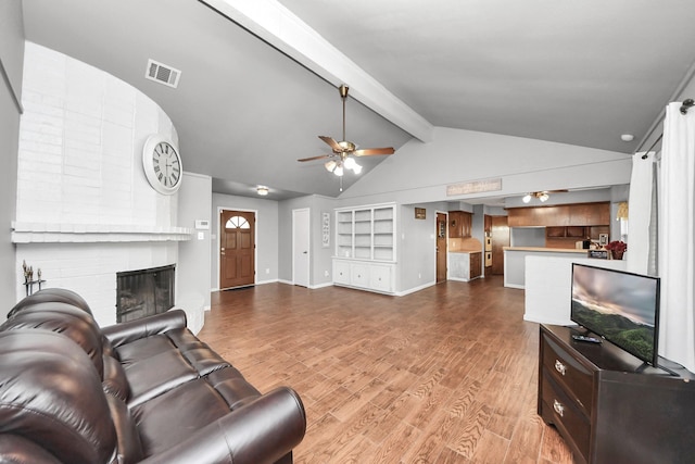 living area with visible vents, a brick fireplace, vaulted ceiling with beams, wood finished floors, and a ceiling fan