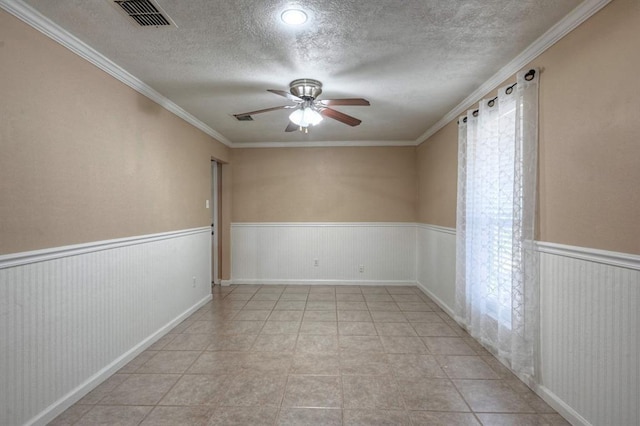 empty room featuring a wainscoted wall, visible vents, ceiling fan, a textured ceiling, and crown molding