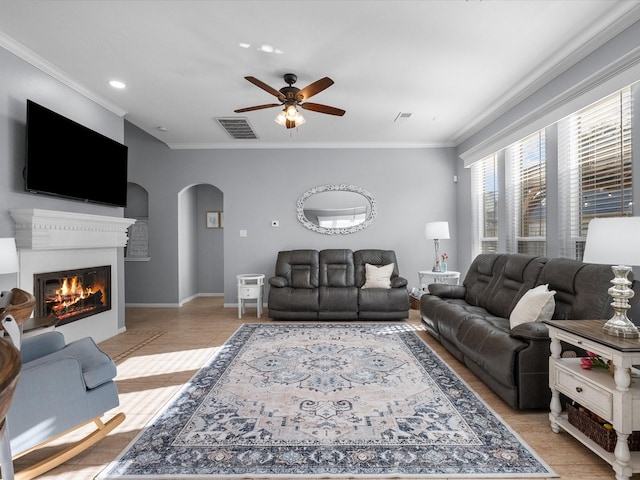 living area featuring visible vents, ornamental molding, a glass covered fireplace, arched walkways, and ceiling fan