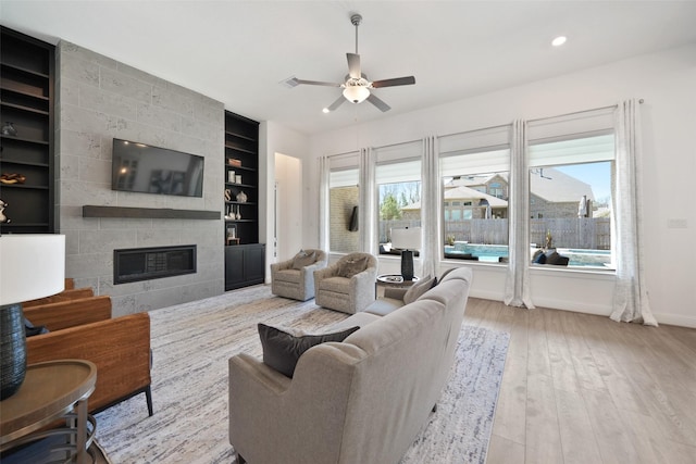 living room featuring a ceiling fan, a tiled fireplace, built in features, wood finished floors, and baseboards