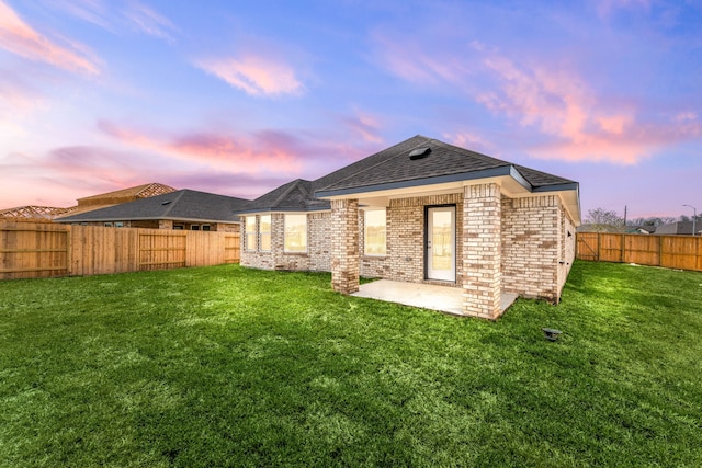 back of house at dusk featuring brick siding, roof with shingles, a fenced backyard, a yard, and a patio