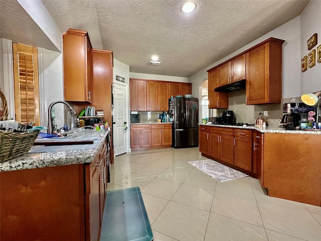 kitchen with light stone counters, freestanding refrigerator, a sink, under cabinet range hood, and brown cabinets