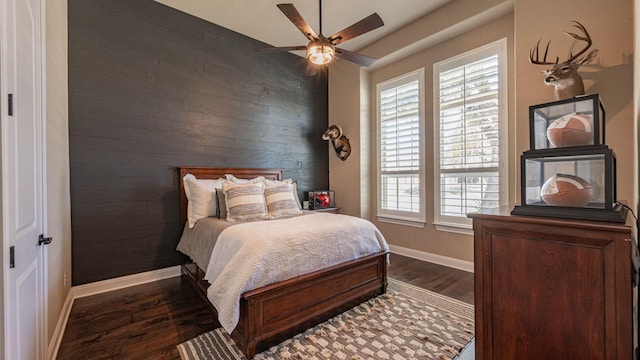 bedroom with wooden walls, an accent wall, ceiling fan, baseboards, and dark wood-style floors