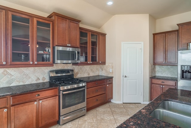 kitchen with dark stone countertops, appliances with stainless steel finishes, light tile patterned floors, glass insert cabinets, and vaulted ceiling