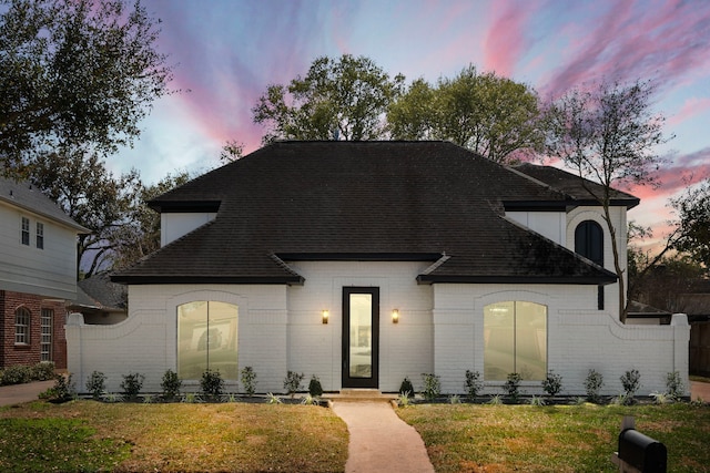 french country style house with a lawn, brick siding, and roof with shingles