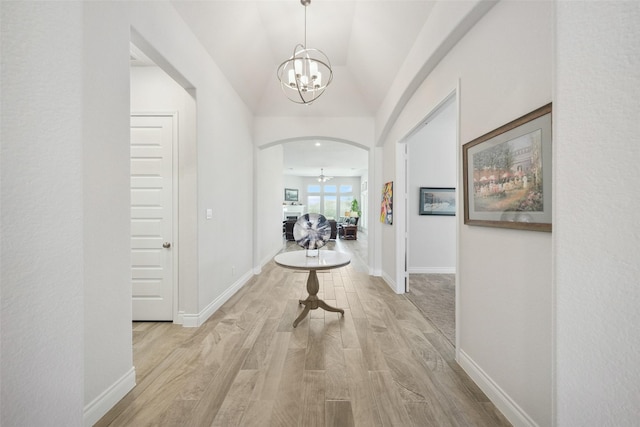 hallway featuring light wood finished floors, baseboards, vaulted ceiling, arched walkways, and a notable chandelier