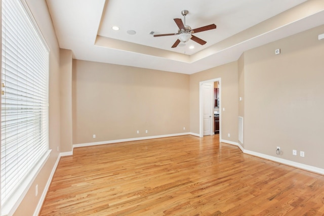 empty room with a healthy amount of sunlight, baseboards, light wood-type flooring, a tray ceiling, and a ceiling fan