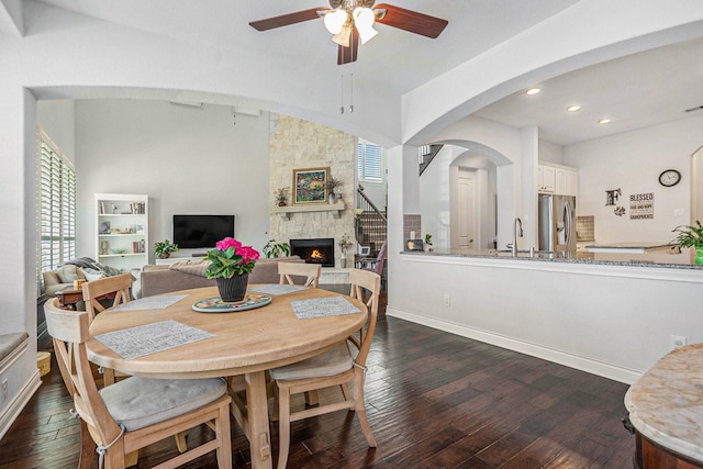 dining area featuring a ceiling fan, a stone fireplace, dark wood-style floors, and baseboards