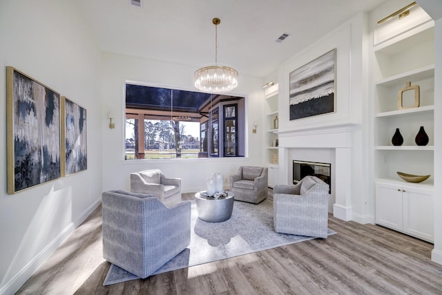 living room featuring built in shelves, wood finished floors, visible vents, a glass covered fireplace, and a chandelier