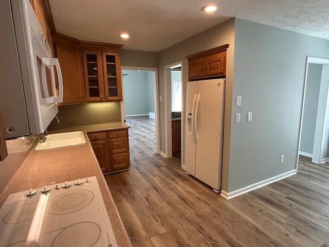 kitchen with white appliances, brown cabinetry, wood finished floors, baseboards, and glass insert cabinets