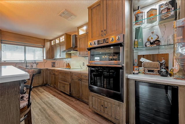 kitchen featuring visible vents, black electric stovetop, wall chimney range hood, beverage cooler, and stainless steel double oven