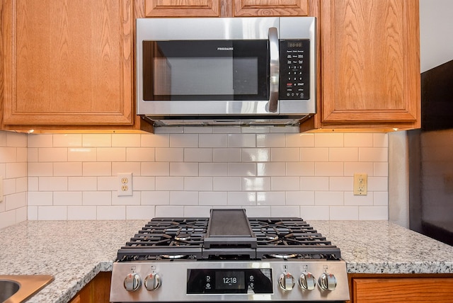 kitchen with tasteful backsplash, stainless steel appliances, brown cabinetry, and light stone countertops