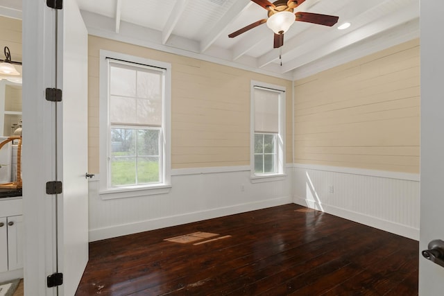 spare room with baseboards, ceiling fan, beamed ceiling, a wainscoted wall, and dark wood-style flooring