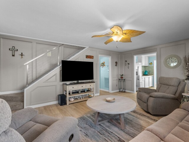 living room featuring a ceiling fan, a decorative wall, stairs, and light wood-style flooring