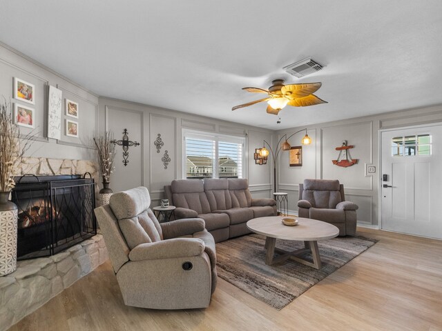 living area featuring wood finished floors, visible vents, a ceiling fan, a stone fireplace, and a decorative wall