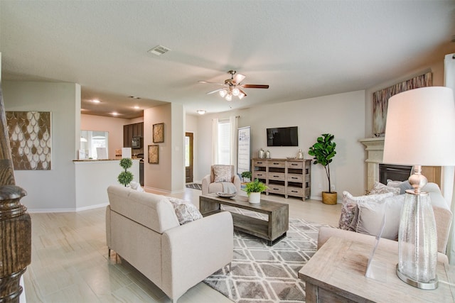 living area featuring visible vents, a ceiling fan, a textured ceiling, a glass covered fireplace, and baseboards