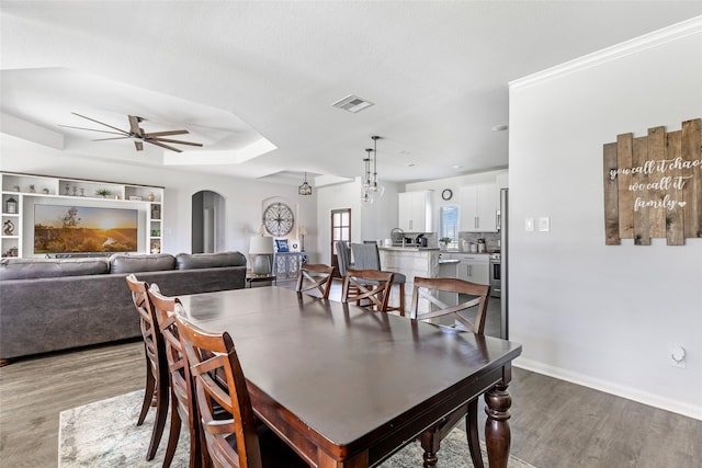 dining space with visible vents, light wood-type flooring, a tray ceiling, arched walkways, and a ceiling fan