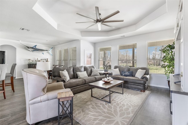 living area featuring plenty of natural light, a ceiling fan, a tray ceiling, and dark wood-style flooring