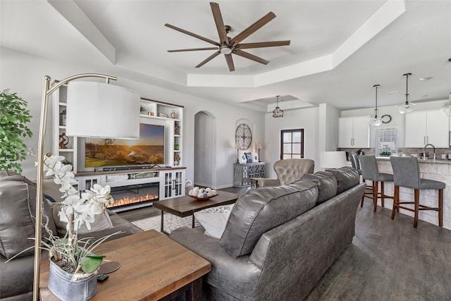 living area featuring ceiling fan, arched walkways, a glass covered fireplace, a raised ceiling, and dark wood-style flooring