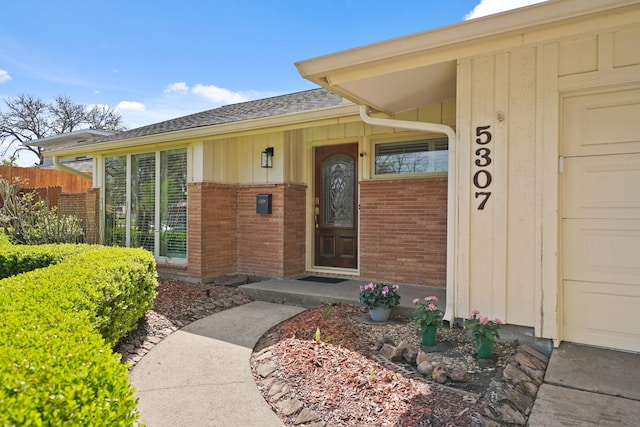 doorway to property featuring brick siding, fence, board and batten siding, and roof with shingles