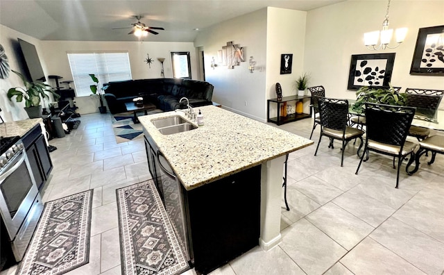 kitchen with an island with sink, ceiling fan with notable chandelier, a sink, light stone counters, and stainless steel range with gas cooktop