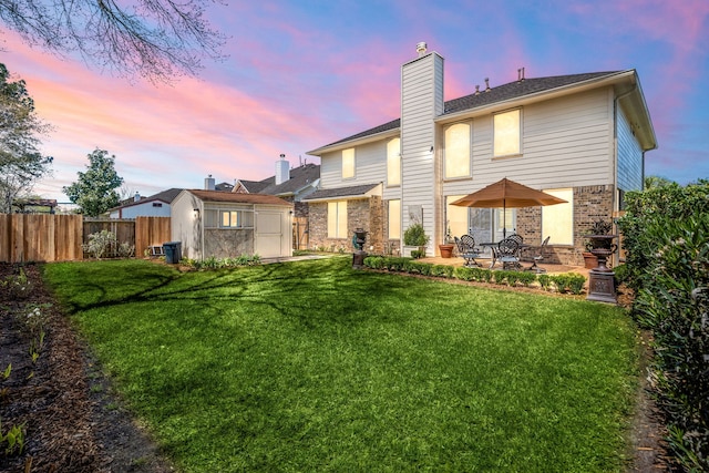 back of house at dusk featuring an outbuilding, fence, a chimney, a patio area, and a lawn