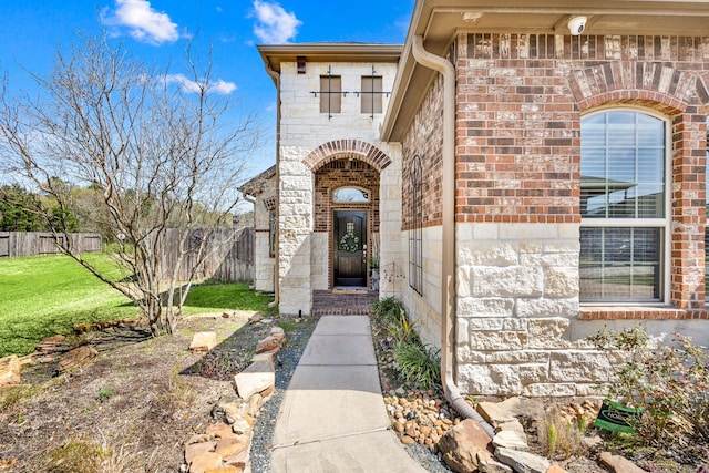 view of exterior entry featuring brick siding, fence, stone siding, and a lawn