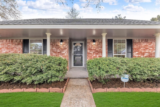 doorway to property with brick siding, covered porch, and roof with shingles